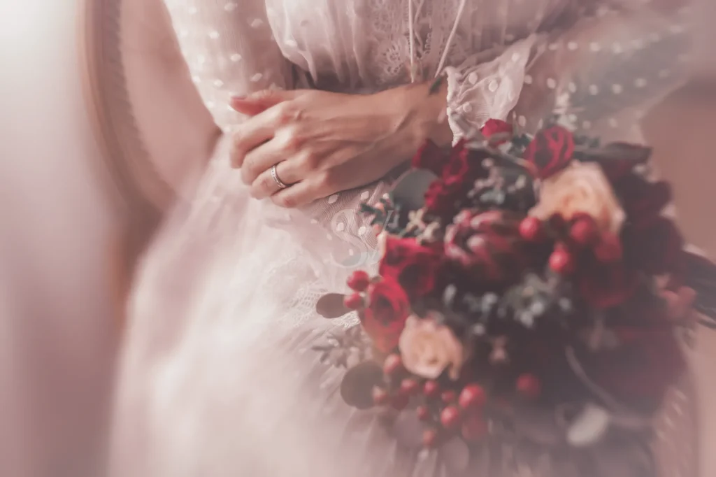 Bride's hand with wedding ring beside a red floral bouquet